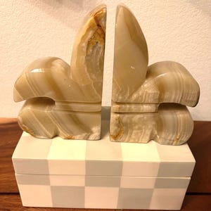 May include: A pair of onyx bookends with a unique, abstract design. The bookends are a light beige color with brown and tan veining, and are set on a white and gray checkered box. These decorative bookends add a touch of elegance to any bookshelf.
