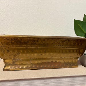 May include: A rectangular, antique-style brass planter with a distressed finish. The planter has a raised base and a decorative dotted pattern along the sides. It sits atop a stack of books, with a plant in the background.