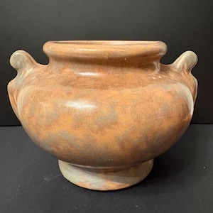 May include: A brown ceramic vase with two small handles on either side. The vase has a wide, rounded body and a slightly flared rim. The base is a lighter shade of brown. The vase is likely handmade and has a rustic appearance.