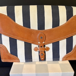 May include: A rectangular clutch bag featuring a navy blue and off-white vertical striped canvas body. The bag is accented with tan leather trim on the corners and a wide, curved leather flap closure secured by a central leather strap and metal stud.