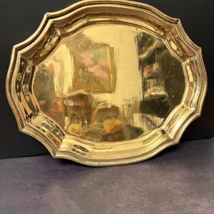 May include: A gold-colored, oval-shaped tray with a scalloped edge. The tray has a reflective surface, and the interior reflects the surrounding environment. The tray is made of metal and has a decorative design.