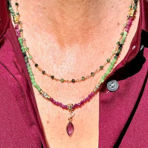 May include: A layered necklace featuring green, pink, and black gemstone beads strung on gold-colored chains. The bottom layer has a pendant with a purple teardrop-shaped stone. The necklace is displayed against a burgundy shirt.