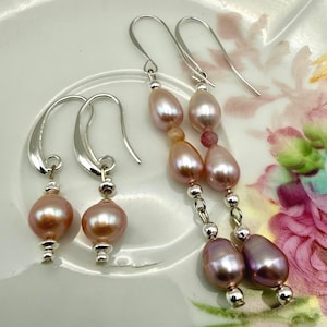 May include: Two pairs of earrings with pink freshwater pearls. One pair features a single pearl drop, while the other has multiple pearls and small beads. The earrings have silver-colored hooks and accents, and are displayed on a white plate.