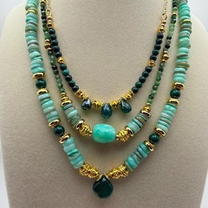 May include: A three-strand necklace set featuring turquoise and green beads with gold-colored accents. The necklaces have various shapes of beads, including round, disc, and teardrop. The necklace is displayed on a mannequin.
