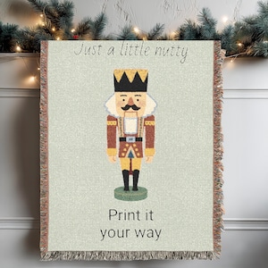 May include: A woven blanket featuring a colorful illustration of a nutcracker soldier with a black crown and mustache. The blanket has the text "Just a little nutty" and "Print it your way". The blanket has a fringed edge.