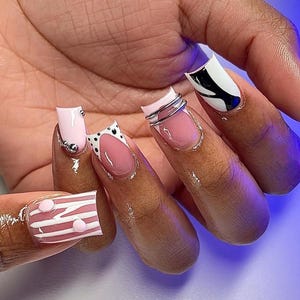May include: A hand displaying a set of manicured nails with various designs. The nails feature pink, white, and black colors, with polka dots, stripes, and metallic accents. Some nails have silver rings attached.