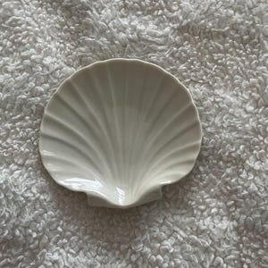 May include: A white ceramic dish shaped like a scallop shell. The dish features a smooth, glossy finish with raised, radiating ridges that extend from the base to the scalloped edge, creating a fan-like appearance with a concave center.