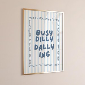 May include: A framed print with a gold-colored frame. The artwork features vertical blue and cream stripes. The text "BUSY DILLY DALLY ING" is written in blue, within a wavy blue border.