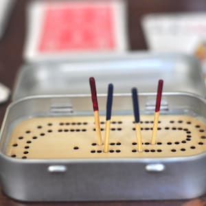 May include: A metal cribbage board with a tan interior and a pattern of holes. Four wooden pegs with red and blue tips are inserted into the board. The board is inside a silver tin.
