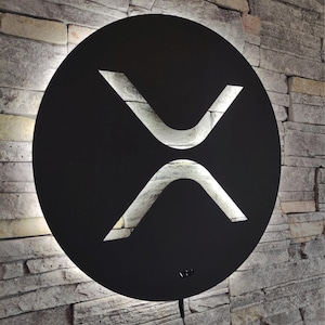 May include: A black, circular wall art piece featuring the XRP symbol, illuminated with a bright, white light. The symbol is cut out of the black circle, revealing the light behind it. The art is mounted on a textured, gray stone wall.