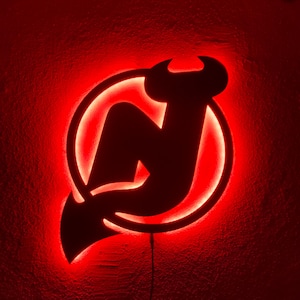 May include: A red illuminated sign displaying the New Jersey Devils logo. The logo is a black silhouette of a stylized "N" with horns and a tail, within a circular border. The sign glows with a red light, creating a dramatic effect.