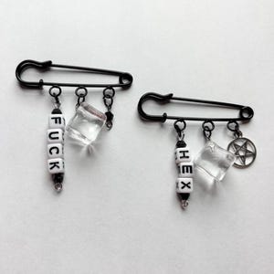 May include: Two black safety pins with dangling charms. One pin has the word "FUCK" spelled out in white letter beads, a clear cube, and a small charm. The other pin has "HEX" spelled out, a clear cube, and a pentagram charm.