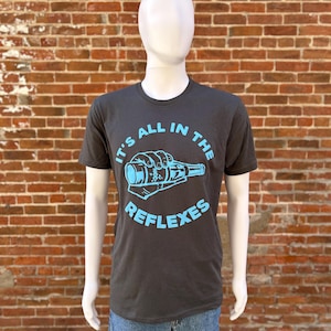 May include: Dark gray t-shirt with the text "IT'S ALL IN THE REFLEXES" in a light blue color. The graphic shows a hand holding a bottle. The shirt is displayed on a mannequin.