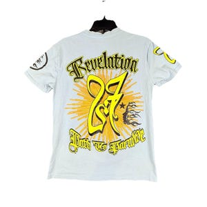 Könnte beinhalten: Hellblaues T-Shirt mit einem Grafikdesign auf der Rückseite. Das Design zeigt das Wort "Revelation" über einer großen gelben "27" mit einem Sonnenstrahlen-Hintergrund. Darunter steht der Text "Path to Paradise".