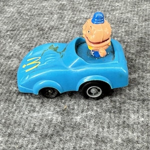 Puede incluir: Un pequeño coche de juguete azul con ruedas negras y un logotipo "M" amarillo en el capó. Un personaje de dibujos animados con un sombrero azul está sentado en el coche. El juguete es de plástico y está sobre una superficie gris.