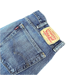May include: Close-up of blue denim jeans with a back pocket and a Levi Strauss & Co. label. The label has red and white text and a red logo. The jeans have a classic five-pocket design.