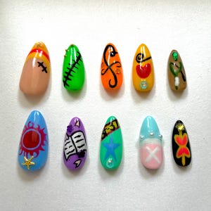 May include: A set of ten artificial nails with various designs. The nails feature colors like blue, green, orange, and pink, with patterns including a sun, a star, and abstract shapes. Some nails have embellishments.