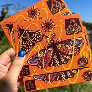May include: A rectangular orange card featuring a repeating print of moths, flowers, and stars. The moths have dark bodies with patterned wings in shades of purple, orange, and yellow. Thin, wavy yellow lines are drawn across the surface.