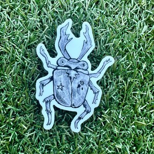 May include: A sticker featuring a detailed illustration of a beetle with large antlers and six legs. The beetle is drawn in black and white with a star and floral design on its back. The sticker is on a white background.