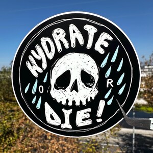 Könnte beinhalten: Runder schwarzer Aufkleber mit weißem Text und einem Totenkopf-Motiv. Die Worte "HYDRATE or DIE!" sind in einem Kreis um einen weißen Totenkopf angeordnet. Hellblaue Wassertropfen befinden sich auf der rechten Seite.