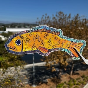 May include: A yellow fish sticker with a glittery border. The fish has a detailed design with orange fins and a blue eye. The sticker is held up against a background of trees and a blue sky.