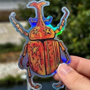 May include: Holographic sticker depicting an orange and yellow beetle with star details. The sticker has a reflective, iridescent finish. The design is held by a hand against a blurred green background. The beetle design is detailed and whimsical.