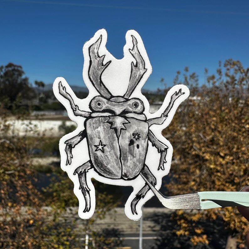 May include: A black and white illustration of a beetle with large antlers, a star, and a floral design. The beetle is held by a pair of tweezers against a backdrop of trees and a blue sky. The beetle is a sticker.