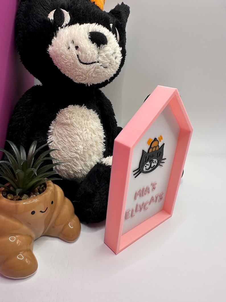 Op de afbeelding: Een roze omlijst bord met de tekst "Mia's Ellycats" en een zwarte kattenafbeelding. Een zwart-witte pluche kat en een bruine, croissantvormige plantenbak met een kleine vetplant staan ook in de afbeelding.