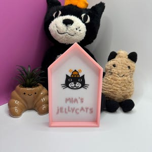 Op de afbeelding: Een roze huisvormig bord met de tekst "MIA'S JELLYCATS" en een zwarte kattenafbeelding. Een zwart kattenknuffel met een oranje hoed staat achter het bord. Een kleine bruine plantenbak met een plant en een beige knuffel staan ook in de afbeelding.