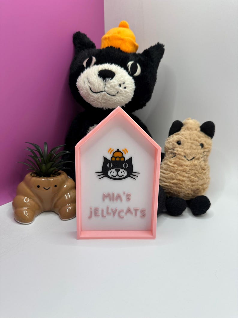 Op de afbeelding: Een roze huisvormig bord met de tekst "MIA'S JELLYCATS" en een zwarte kattenafbeelding. Een zwart kattenknuffel met een oranje hoed staat achter het bord. Een kleine bruine plantenbak met een plant en een beige knuffel staan ook in de afbeelding.