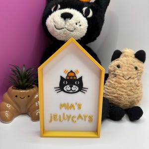 Op de afbeelding: Een geel omrand bord met de tekst "MIA'S JELLYCATS" en een zwarte kattenafbeelding met een hoed. Een zwart kattenknuffel met een oranje hoed staat achter het bord. Een kleine vetplant in een bruine pot en een bruin knuffelbeest zijn ook zichtbaar.