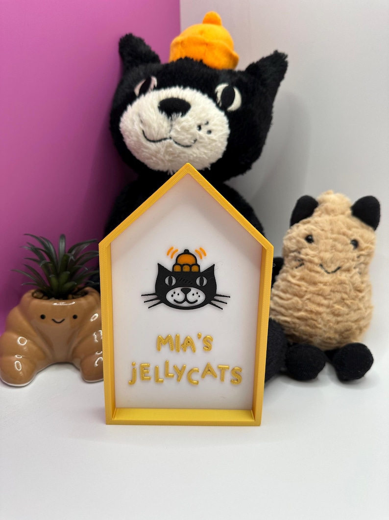 Op de afbeelding: Een geel omrand bord met de tekst "MIA'S JELLYCATS" en een zwarte kattenafbeelding met een hoed. Een zwart kattenknuffel met een oranje hoed staat achter het bord. Een kleine vetplant in een bruine pot en een bruin knuffelbeest zijn ook zichtbaar.