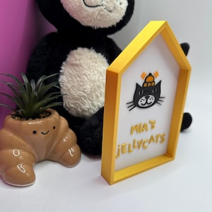 Op de afbeelding: Een geel omrand bord met de tekst "Mia's Jellycats" en een zwarte kattenafbeelding. Een zwart-witte pluche kat en een kleine bruine, croissantvormige plantenbak met een plant staan op de achtergrond.