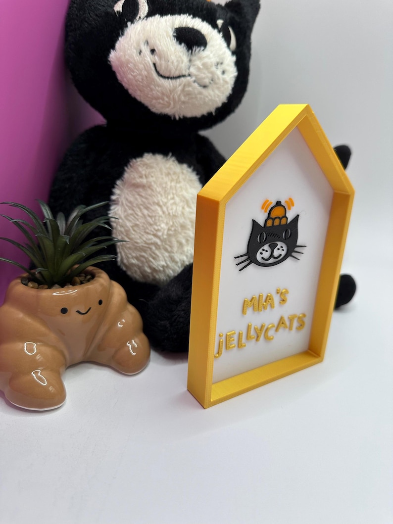 Op de afbeelding: Een geel omrand bord met de tekst "Mia's Jellycats" en een zwarte kattenafbeelding. Een zwart-witte pluche kat en een kleine bruine, croissantvormige plantenbak met een plant staan op de achtergrond.