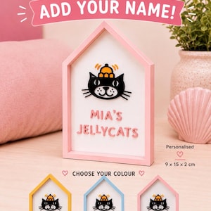 Op de afbeelding: Een roze, huisvormig gepersonaliseerd bord met een illustratie van een zwarte kat met een hoed. Het bord leest "MIA'S JELLYCATS" en "ADD YOUR NAME!". Het bord is 9 x 15 x 2 cm groot.