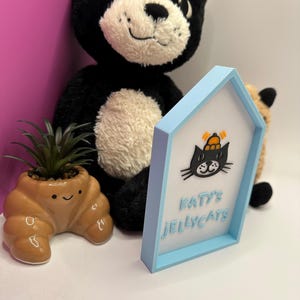 Op de afbeelding: Een zwarte pluche kat met een cr&egrave;mekleurig gezicht en een kleine oranje hoed. Een lichtblauw omlijst bord met de tekst "Katy's Jellycats" en een cartoon kat. Een kleine bruine croissantvormige plantenbak met een groene plant.