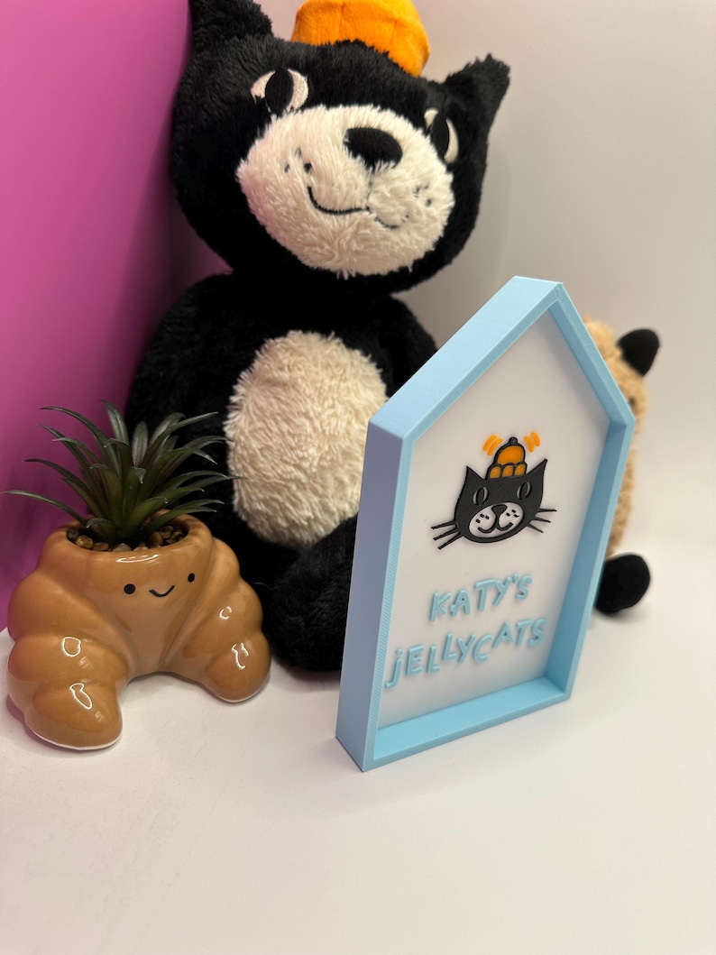 Op de afbeelding: Een zwarte pluche kat met een cr&egrave;mekleurig gezicht en een kleine oranje hoed. Een lichtblauw omlijst bord met de tekst "Katy's Jellycats" en een cartoon kat. Een kleine bruine croissantvormige plantenbak met een groene plant.