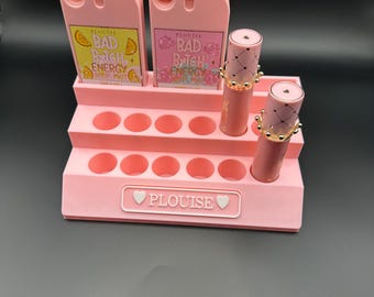 Könnte beinhalten: Ein rosa Make-up-Organizer mit dem Markennamen "PLouise". Er enthält zwei "BAD B*TCH ENERGY" Körpersprays und zwei Lippenstifte mit kronenförmigen Kappen. Die Körpersprays haben Fruchtdarstellungen. Der Organizer hat mehrere kreisförmige Schlitze.