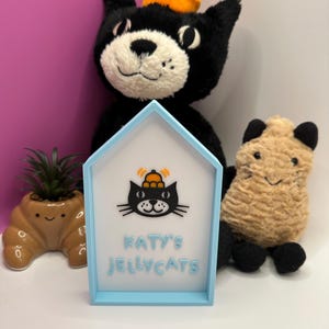 Op de afbeelding: Een lichtblauw huisvormig bord met de tekst "KATY'S JELLYCATS" en een zwarte kattenafbeelding. Een zwart kattenknuffel met een oranje hoed staat achter het bord. Een kleine bruine plantenbak met een vetplant en een bruin knuffelbeest staan ook in de afbeelding.