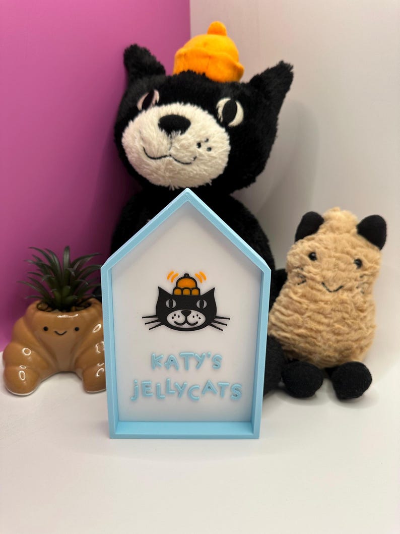 Op de afbeelding: Een lichtblauw huisvormig bord met de tekst "KATY'S JELLYCATS" en een zwarte kattenafbeelding. Een zwart kattenknuffel met een oranje hoed staat achter het bord. Een kleine bruine plantenbak met een vetplant en een bruin knuffelbeest staan ook in de afbeelding.