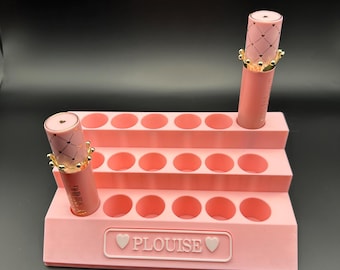 Könnte beinhalten: Ein rosa mehrstufiger Lippenstift-Organizer mit mehreren kreisförmigen Schlitzen. Zwei Lippenstifte mit kronenförmigen Kappen werden gezeigt. Der Organizer hat den Text "PLouise" mit Herzsymbolen. Der Hintergrund ist schwarz.