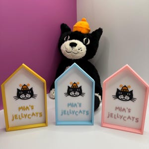 Op de afbeelding: Drie huisvormige frames in geel, blauw en roze, elk met een kattenafbeelding en de tekst "Mia's Jellycats". Een zwart kattenknuffel met een oranje hoed staat op de achtergrond.