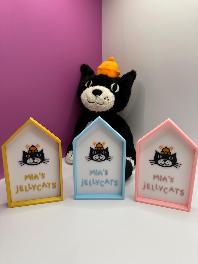 Op de afbeelding: Drie huisvormige frames in geel, blauw en roze, elk met een kattenafbeelding en de tekst "Mia's Jellycats". Een zwart kattenknuffel met een oranje hoed staat op de achtergrond.
