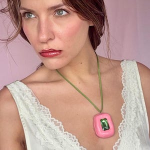May include: A pink pendant necklace with a green gemstone center. The pendant is rectangular with rounded edges and hangs from a green cord. The woman in the image is wearing a white lace-trimmed top.