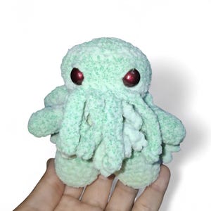 May include: A small, plush, mint-green octopus with red eyes. The tentacles and body are made of a soft, textured material. The toy is held in a hand against a white background. A unique, handmade item.