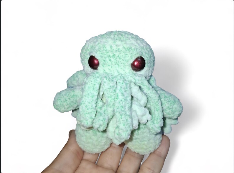 May include: A small, plush, mint-green octopus with red eyes. The tentacles and body are made of a soft, textured material. The toy is held in a hand against a white background. A unique, handmade item.