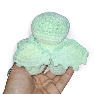 May include: A handmade, light green and white crocheted octopus plush toy. The plush has a round head and eight tentacles. The toy is held in a person's hand against a white background. The octopus is made of a soft, textured yarn.