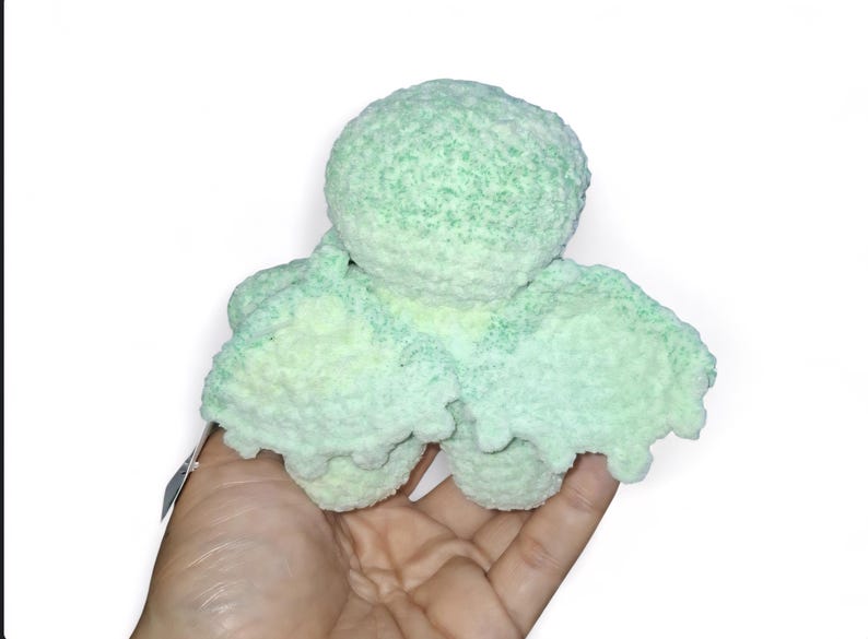 May include: A handmade, light green and white crocheted octopus plush toy. The plush has a round head and eight tentacles. The toy is held in a person's hand against a white background. The octopus is made of a soft, textured yarn.