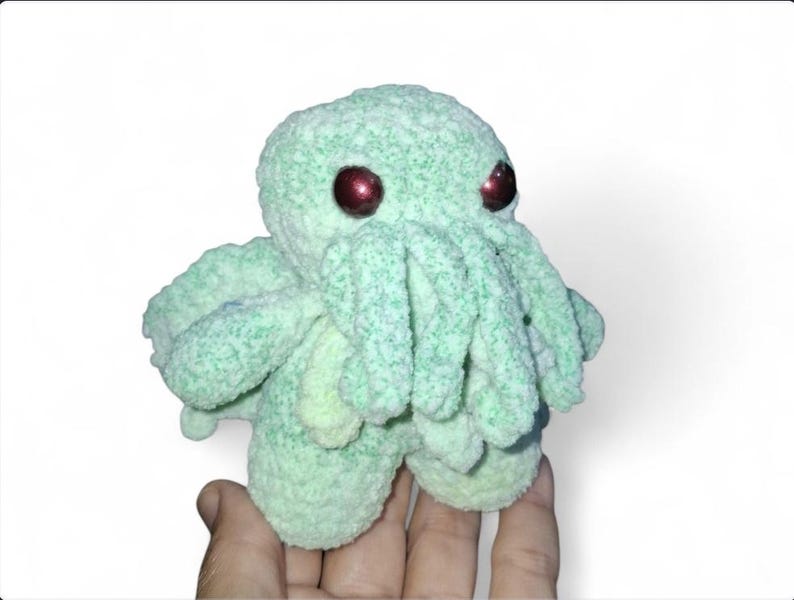 May include: A light green, plush Cthulhu doll with large, red eyes. The handmade creature has multiple tentacles and a textured, fuzzy appearance. The doll is a unique, whimsical item.