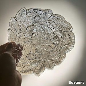 May include: Clear glass decorative bowl with a detailed embossed design of leaves and berries. The bowl has a scalloped edge and is held by a hand. The design is visible in the glass.